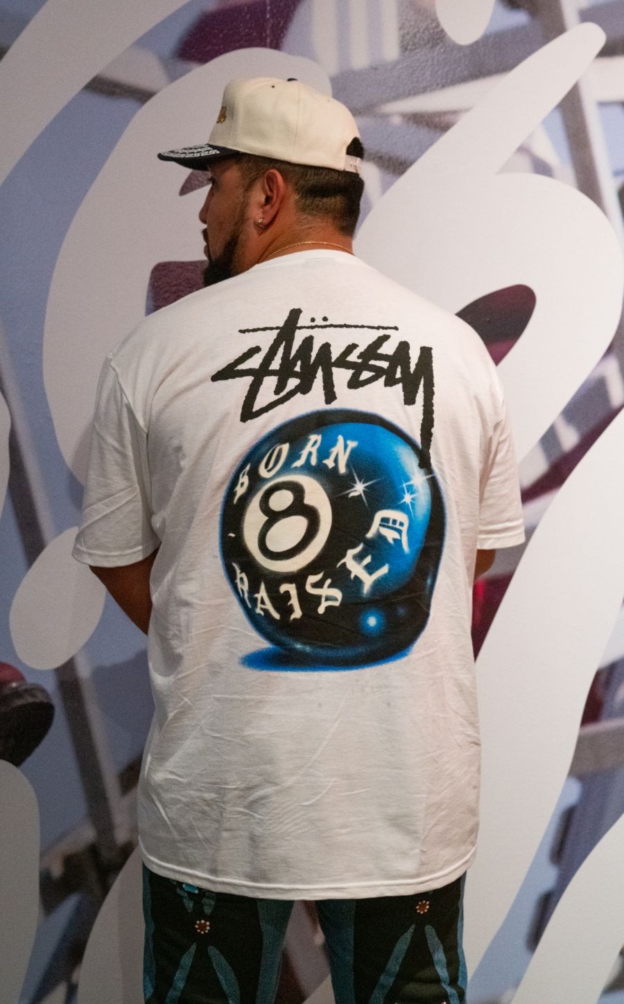 Back view of man standing facing gallery wall. Back of shirt says Stussy Born 8 Raised on an eight ball