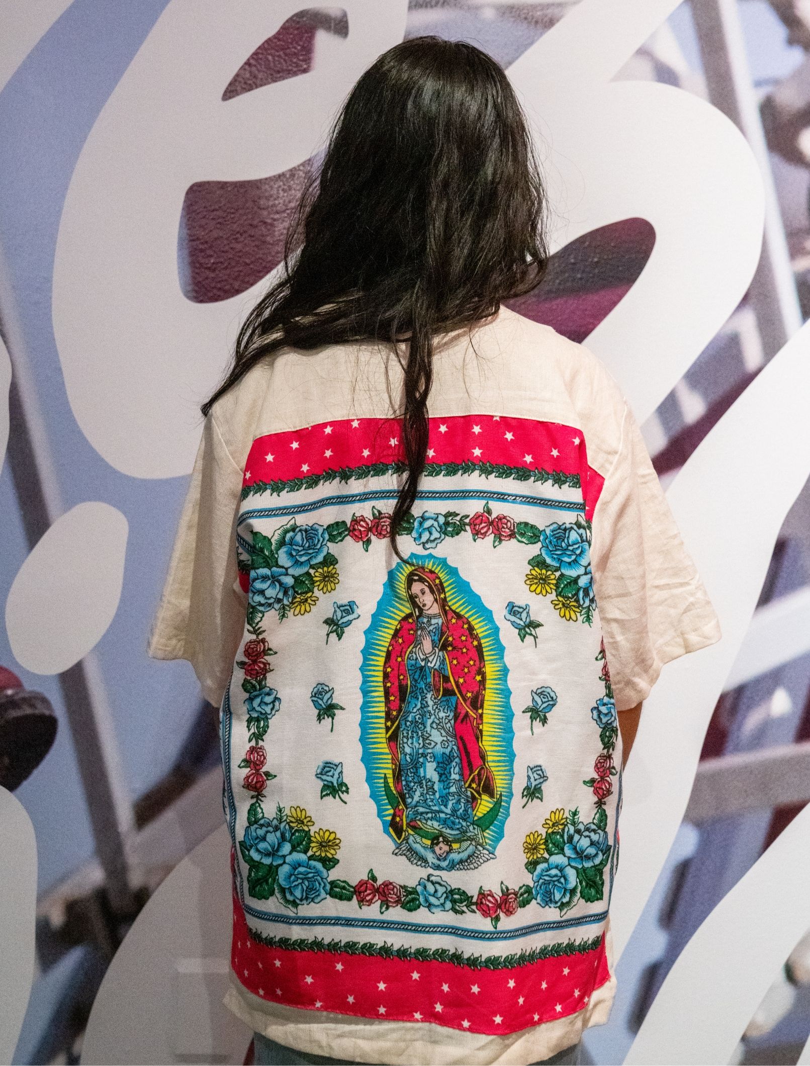 Back view of person posing facing gallery wall. Back of shirt has Mother Mary with roses surrounding her