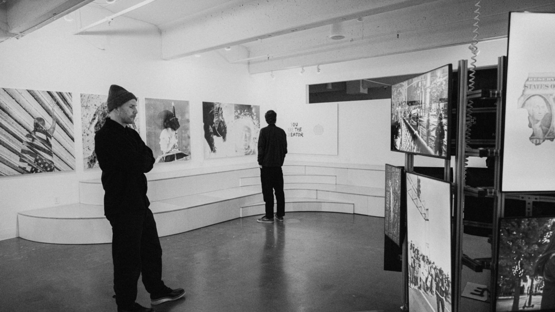 1 person looking at video installation in gallery. A second looks at prints on the wall