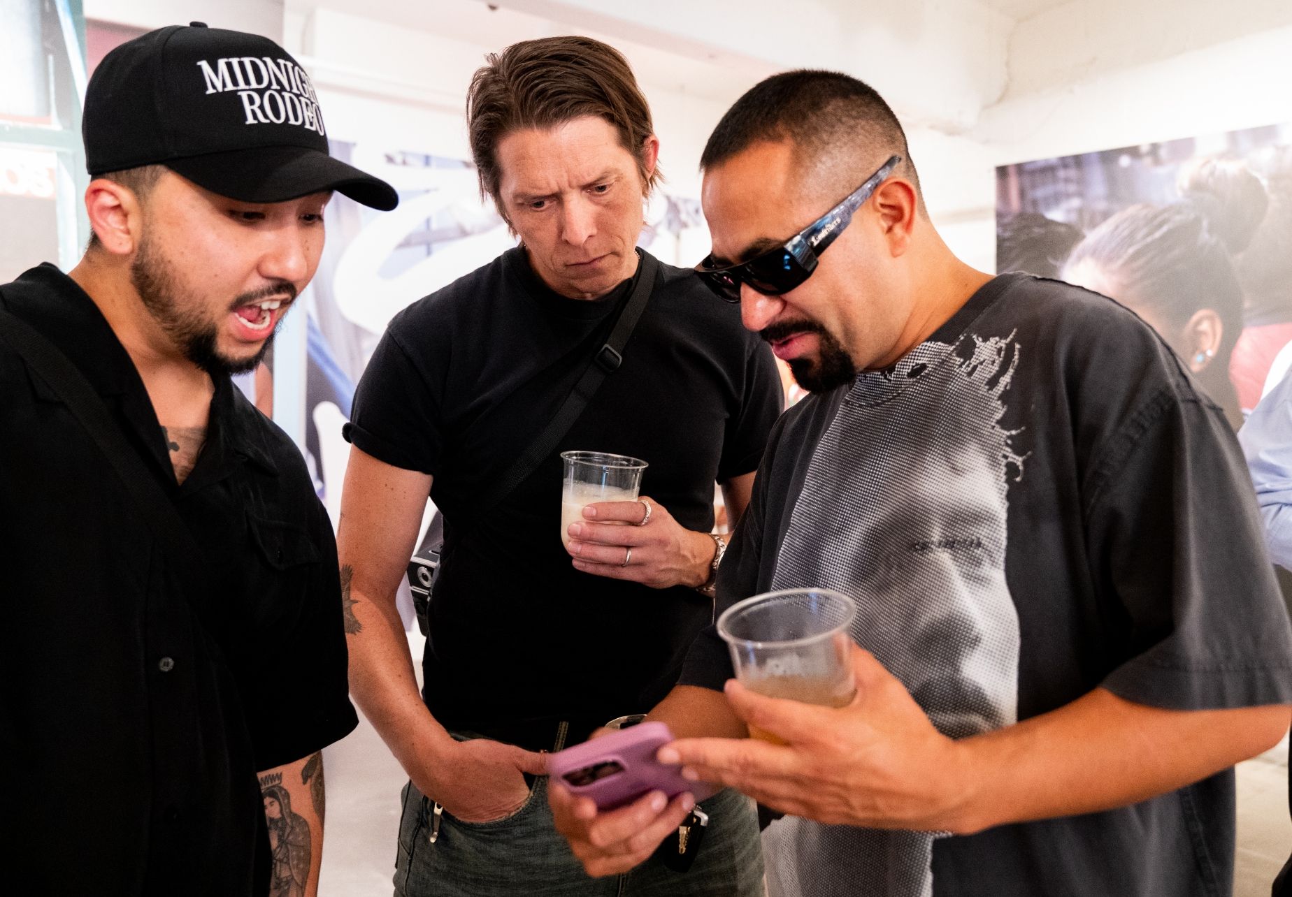 Dorina Lopez shows two gallery show attendees something on his phone. One man has mouth open and the other two have drinks in their hand