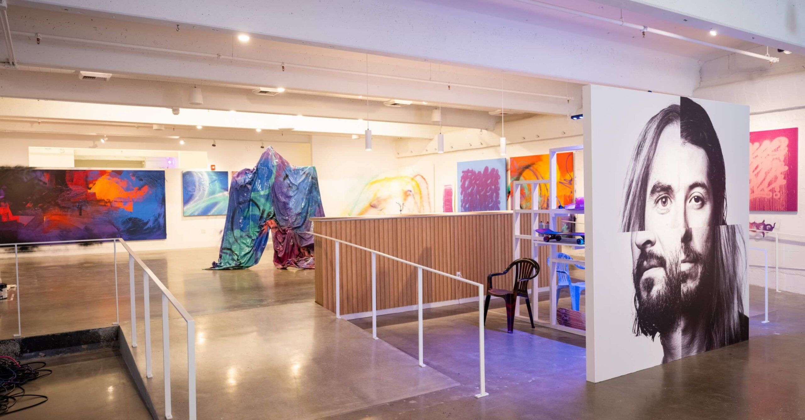 Sentiments Art show in Industry One gallery. Several paintings line 3 walls. A montage of two artists' faces is in center of gallery. Video screens on left wall. Tarp sculpture in foreground.