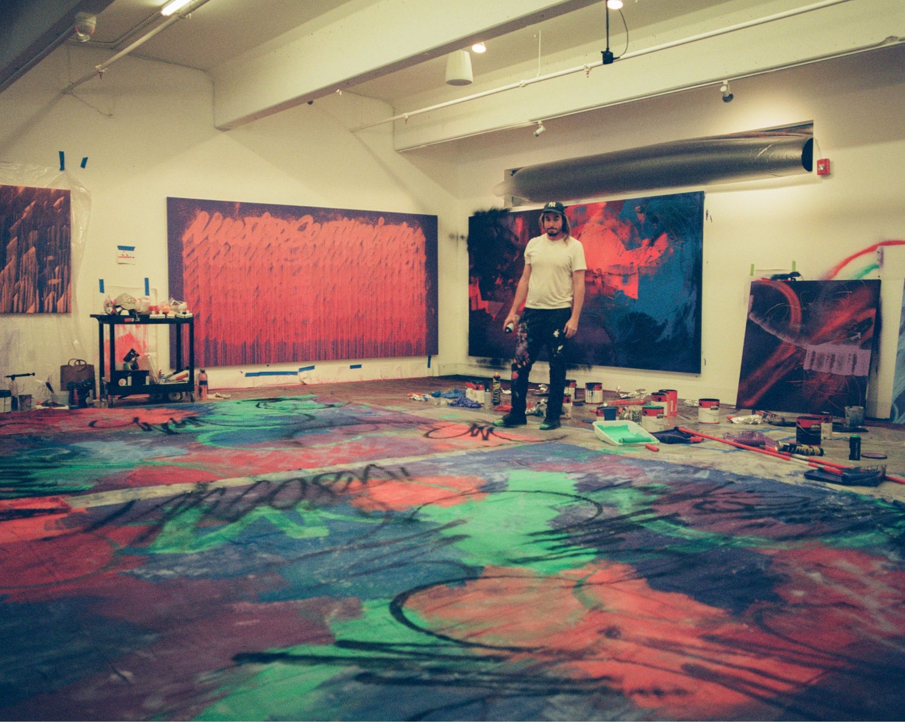 Mad Steez stands in the middle of gallery space with four canvases on wall in progress of being painted. Painted tarps cover the ground along with many buckets of paint, rollers and spray paint cans