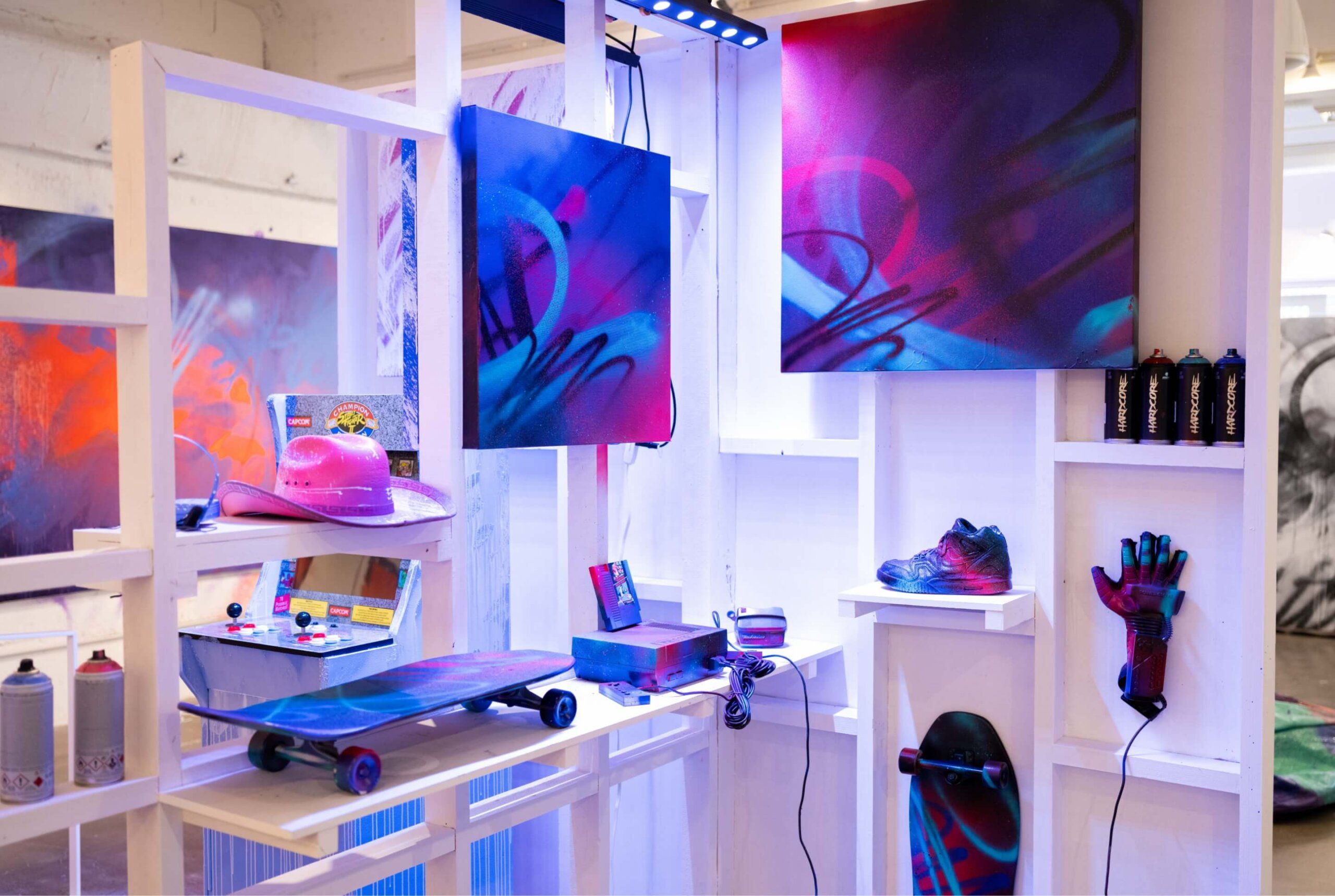 View of spray painted retro items in middlo of art show featuring skateboards, NES, Nintendo power glove and vintage Nike sneaker. Painted canvases line wall behind