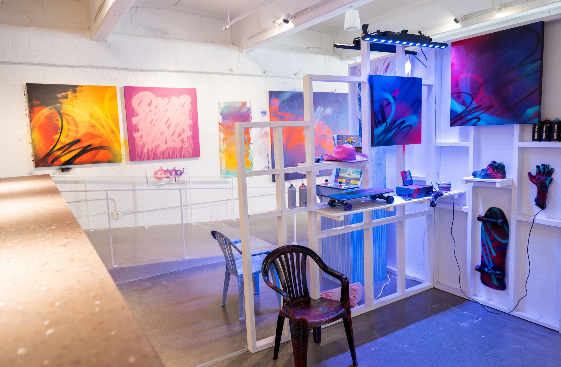 View of spray painted retro items in middlo of art show featuring skateboards, NES, Nintendo power glove and vintage Nike sneaker. Painted canvases line wall behind