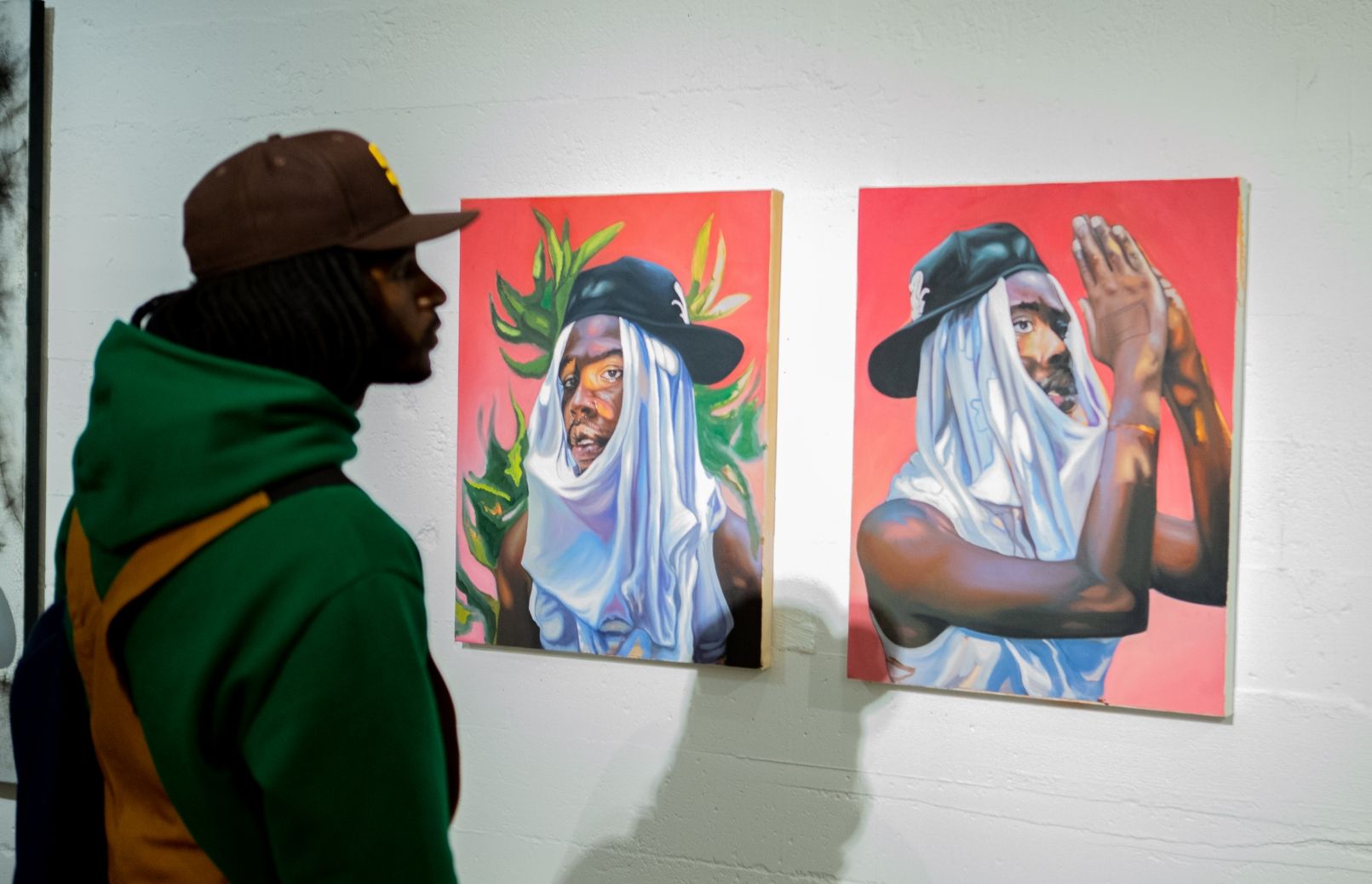 Man looks at two paintings on gallery wall of man with t-sirt and baseball hat on his head